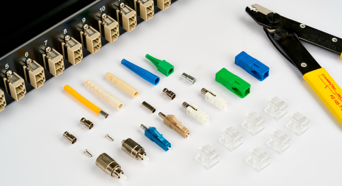 Verschiedene Glasfaser-Stecker und Kupplungen vor einem Patchpanel mit nummerierten Ports.