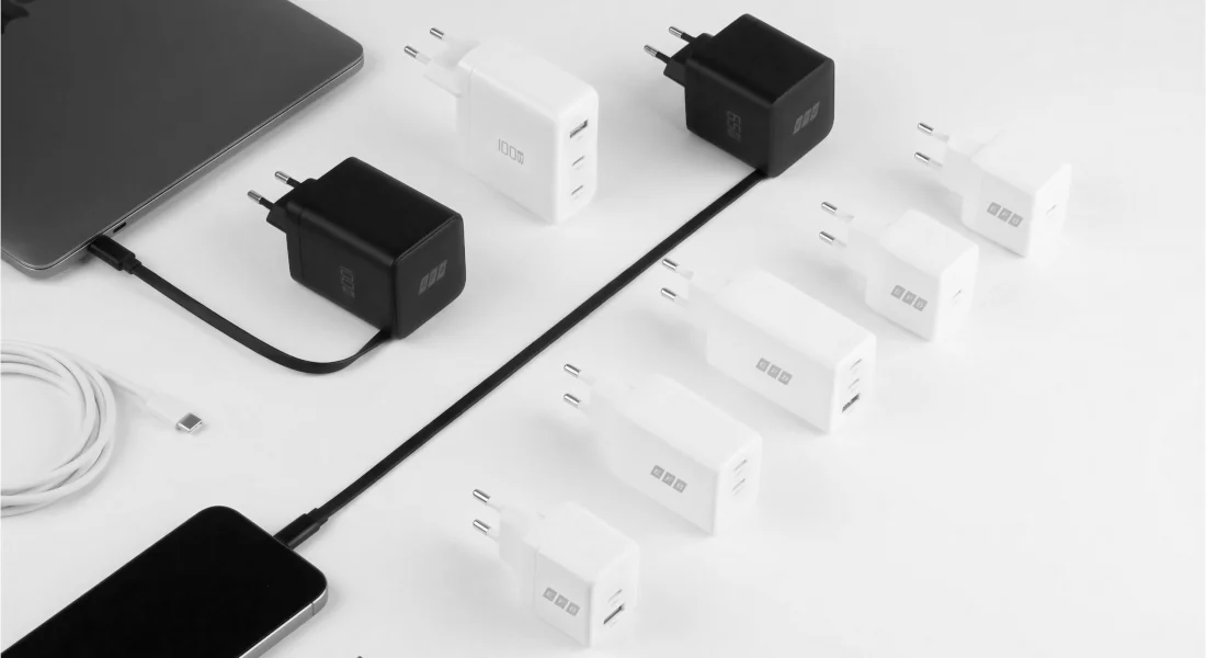 Flere USB-C-opladere i sort og hvid med 65 W og 100 W på hvid baggrund, ved siden af en smartphone, en tablet og et USB-C-opladerkabel