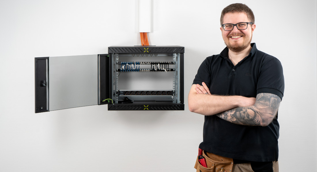 Installatør foran monteret X-Rack 19"-vægkabinet på væggen – dør åben, komponenter overskueligt installeret, ideelt til struktureret netværkskabelføring og hurtig serviceadgang.