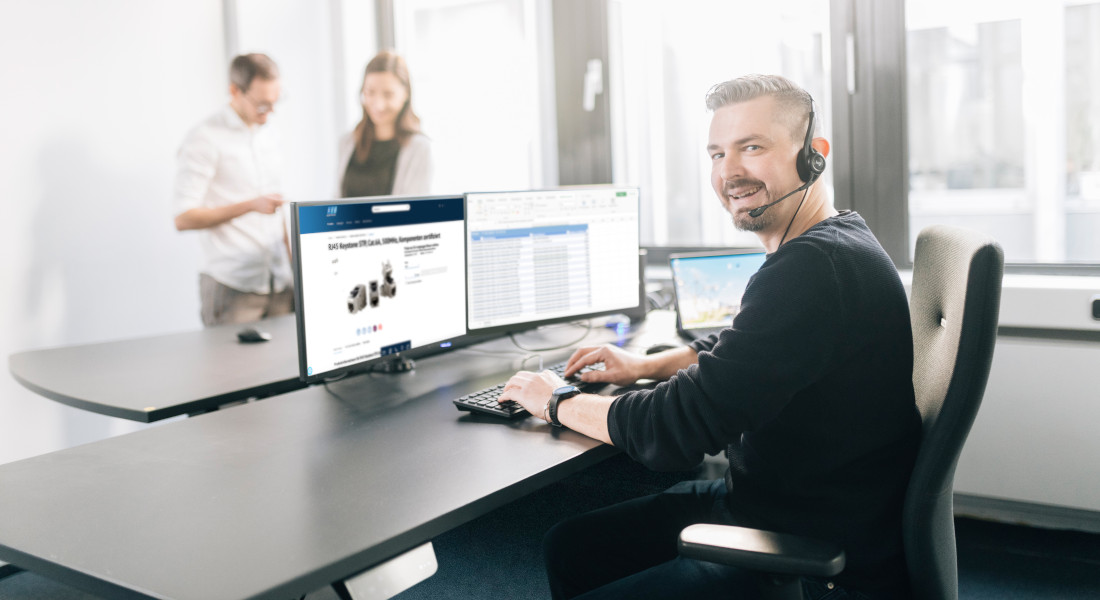 Employee with headset at a desk with two monitors, two people talking in the background