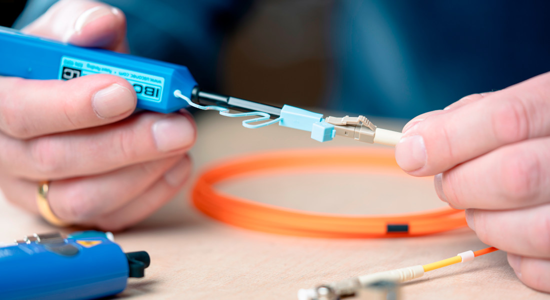 Nahaufnahme einer Person, die ein Glasfaserkabel mit einem Reinigungswerkzeug reinigt, im Hintergrund weitere Glasfaserkabel und ein Adapter. Nahaufnahme einer Person, die ein Glasfaserkabel mit einem Reinigungswerkzeug reinigt, im Hintergrund weitere Glasfaserkabel und ein Adapter.